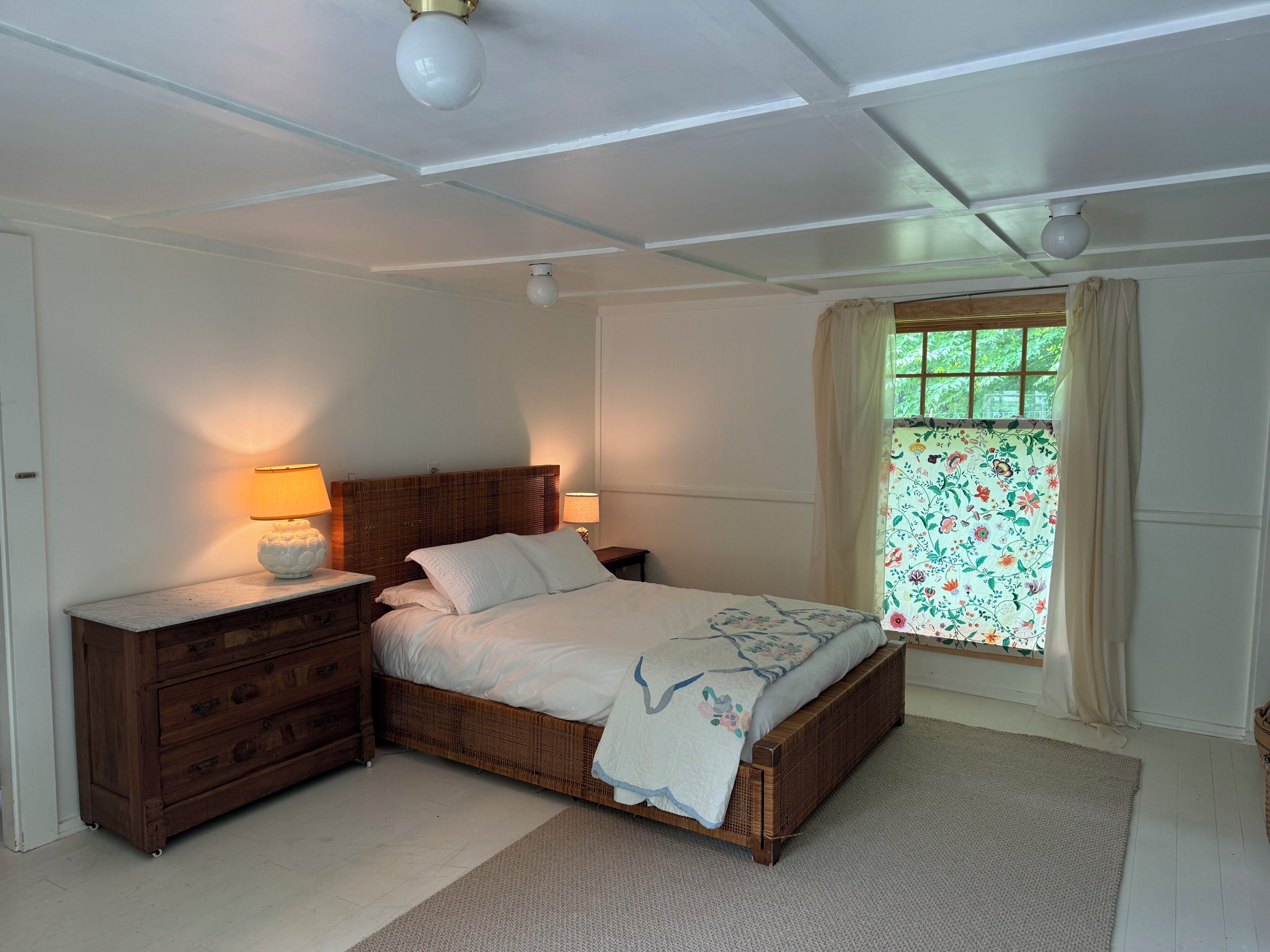Un dormitorio con una cama, dos mesitas de noche con lámparas, un aparador de madera, un ventanal con cortinas florales y un ventilador de techo.