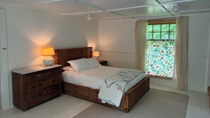 Een slaapkamer met een bed, twee nachtkastjes met lampen, een houten dressoir, een raam met bloemen gordijnen en een plafondventilator.