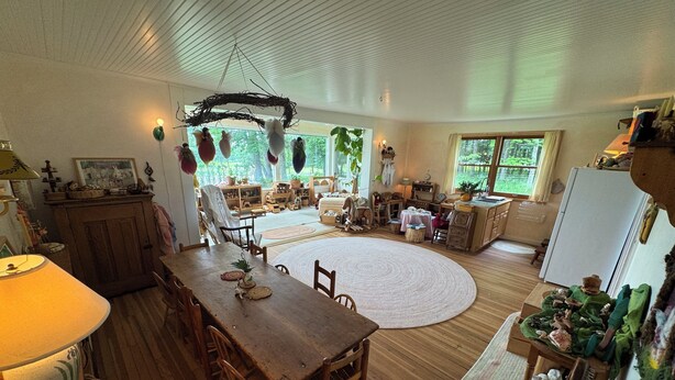 Een ruime kamer met een houten eettafel, een groot rond tapijt en uitzicht op het omliggende landschap door een raam.