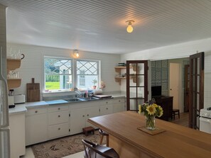 Een keuken met witte kasten, een houten eettafel, een vaas met bloemen en een raam met uitzicht op groen.