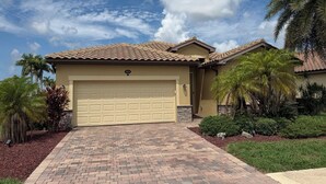 A residential house with a garage, a paved driveway, and landscaped garden with palm trees. - Gated Tuscan Paradise: 4BR/3BA Home + Lagoon Pool, Tennis & Fitness Club Access (Venice)