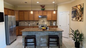 A kitchen with granite countertops, wooden cabinets, a stainless steel refrigerator, a built-in oven, and a stone island with two barstools. - Gated Tuscan Paradise: 4BR/3BA Home + Lagoon Pool, Tennis & Fitness Club Access (Venice)