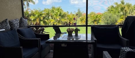 A balcony with wicker furniture, a glass table, and a view of palm trees and a clear sky.