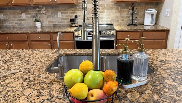 A kitchen with granite countertops, a stainless steel sink, wooden cabinets, and a fruit basket. - Gated Tuscan Paradise: 4BR/3BA Home + Lagoon Pool, Tennis & Fitness Club Access (Venice)