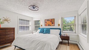 Une chambre à coucher comprenant un lit, une commode, une table de chevet, un ventilateur de plafond et une fenêtre avec des stores.
