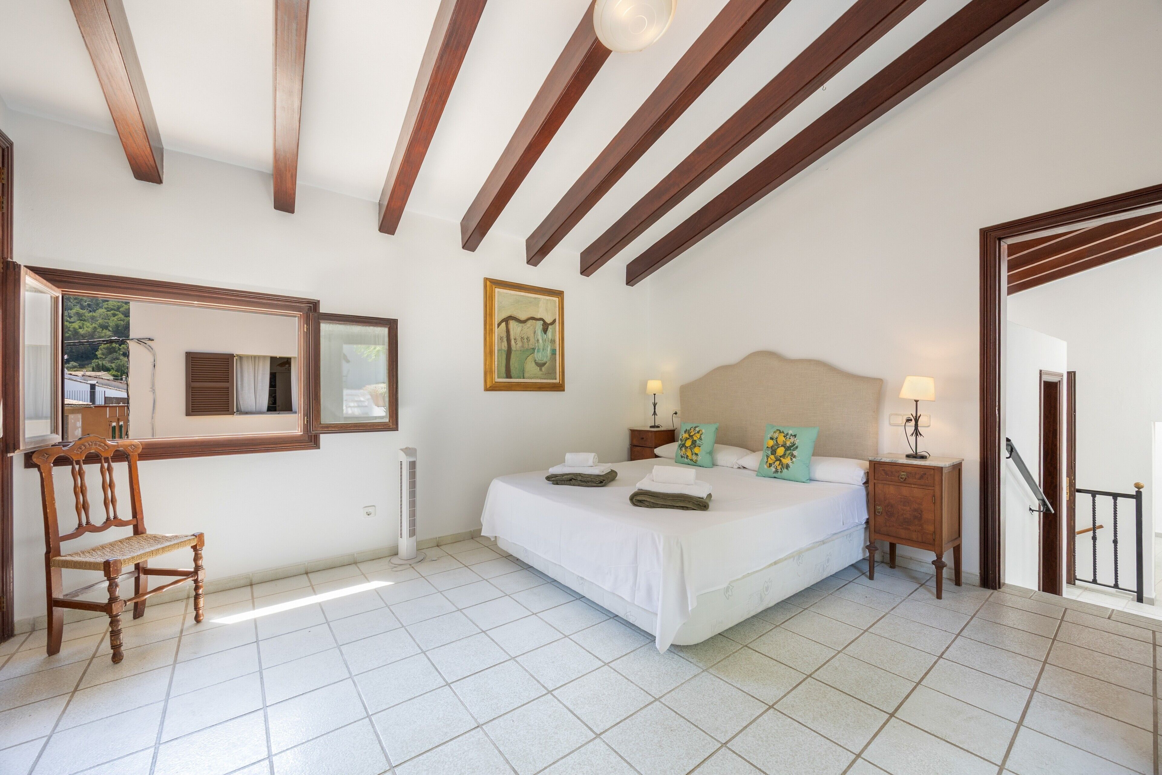 Un dormitorio con cama, mesitas de noche, una silla, una ventana y una puerta.