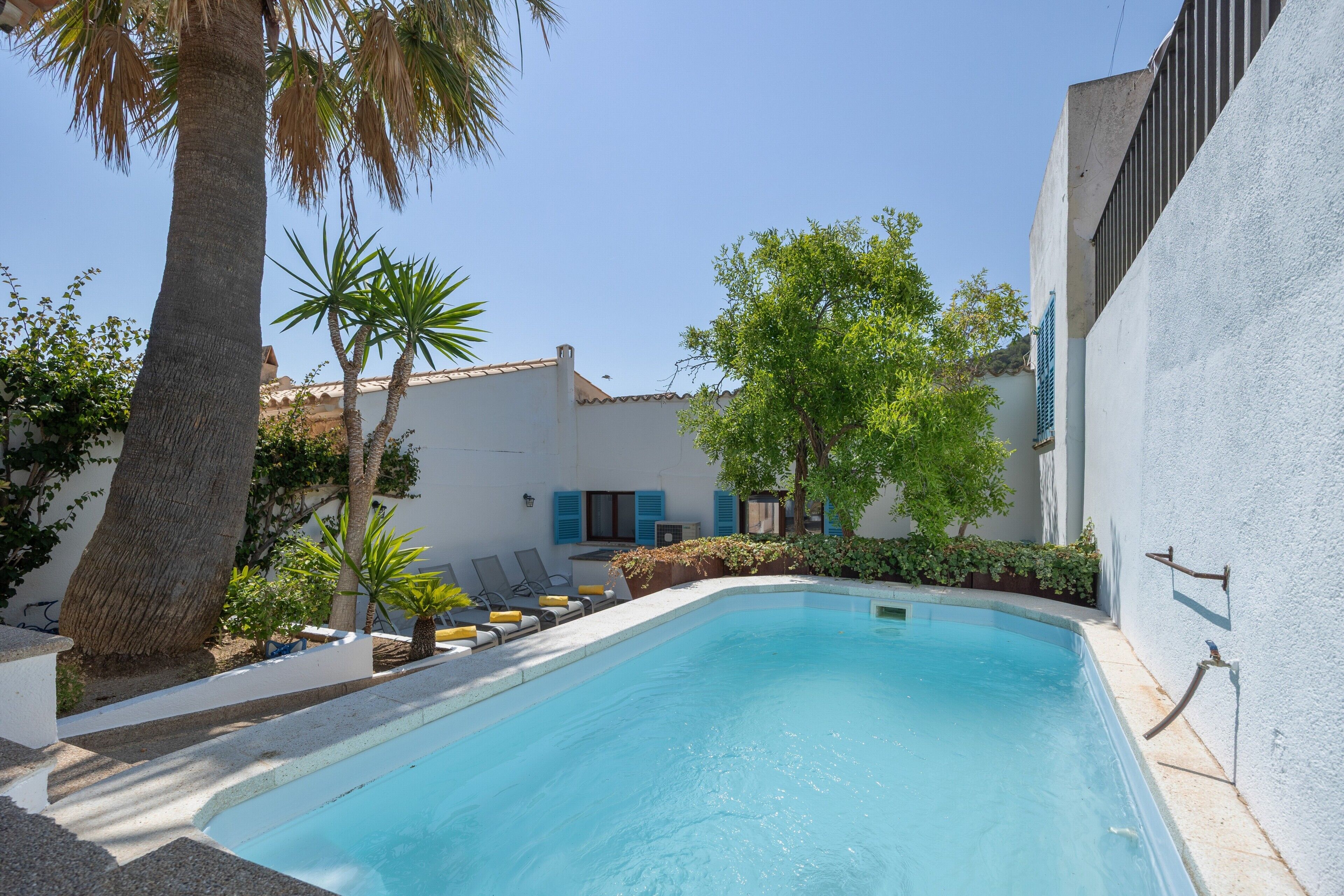 Una piscina con sillones de descanso, palmeras y un edificio blanco con contraventanas azules.
