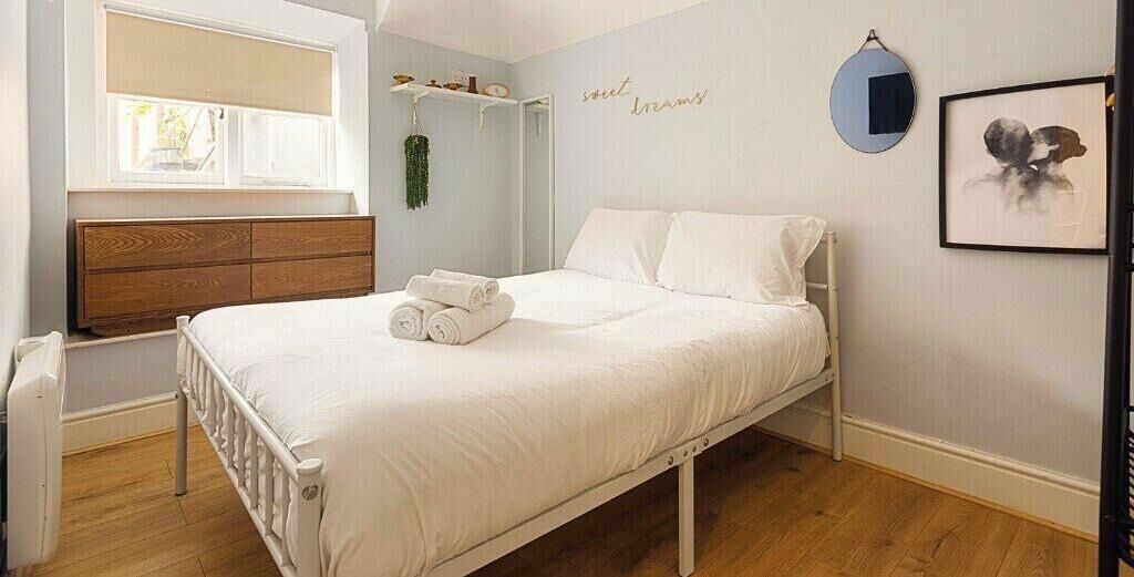 Een slaapkamer met een bed, een raam, een nachtkastje, een wandplank en een ingelijst kunstwerk.