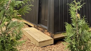 Una cabaña pequeña y oscura con un porche de madera, rodeada de árboles.