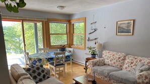 Ein Wohnzimmer mit einer Couch, einem Couchtisch, einem blumengemusterten Sofa, einem Esstisch mit Stühlen und großen Fenstern mit Blick auf Bäume.