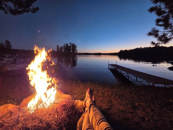 Une personne se détend près d’un feu de camp, avec un lac et une jetée en arrière-plan.