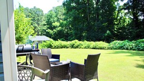 Een terras buiten met rieten meubels, een barbecue en uitzicht op een verzorgd gazon en bomen.