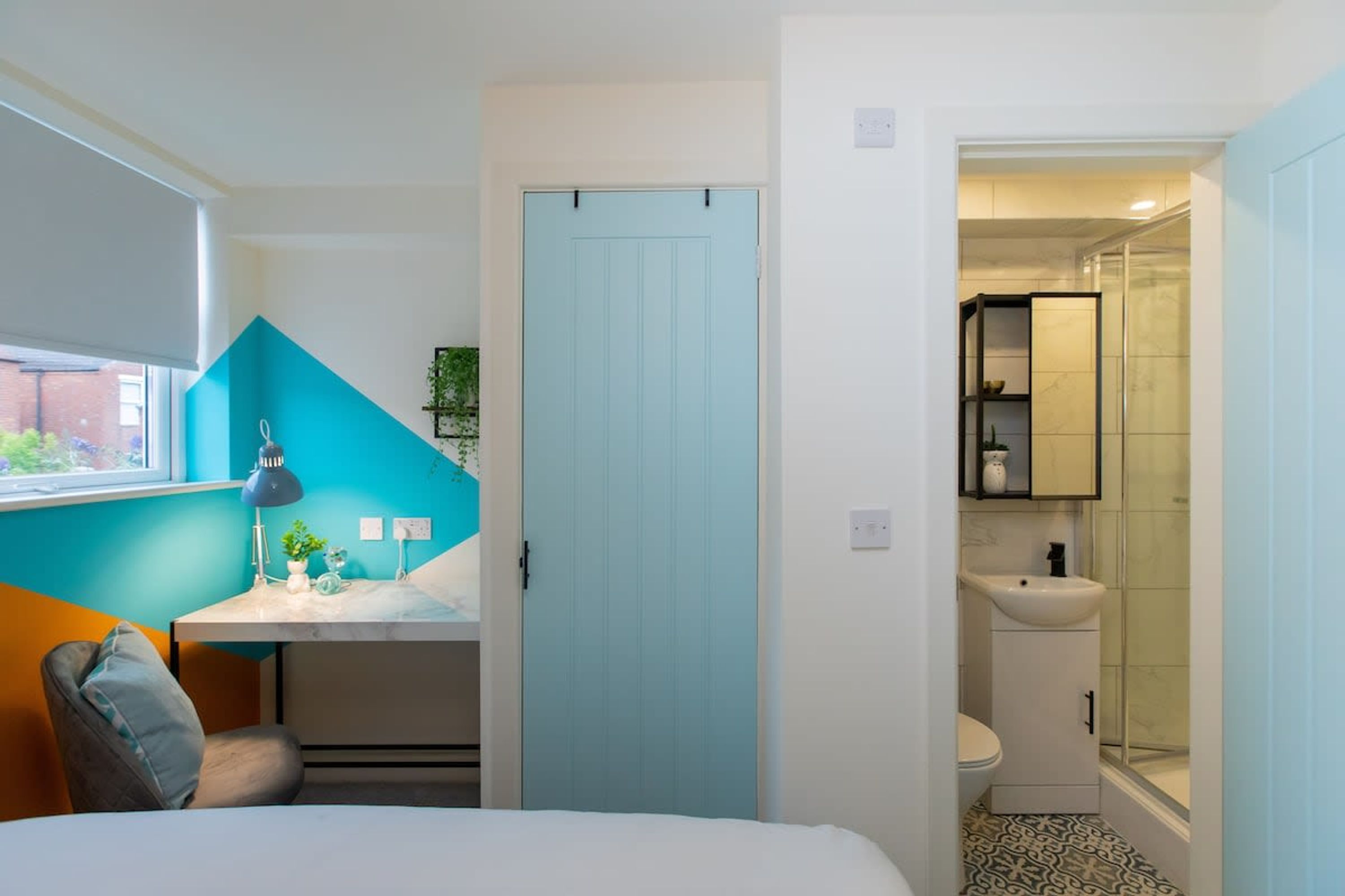 Ein Schlafzimmer mit Bett, Schreibtisch, Stuhl, einer blauen Tür und einem Badezimmer mit Waschbecken und Dusche.