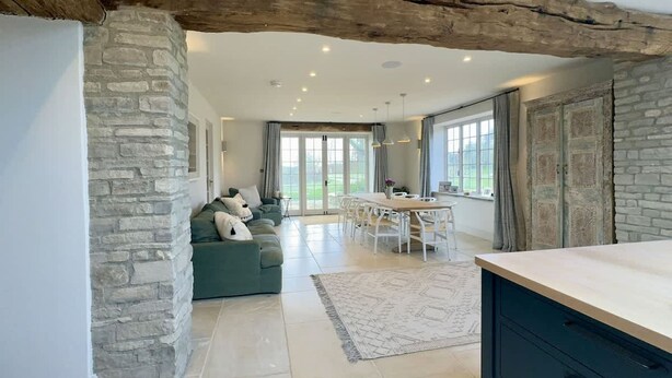 A spacious living area with a stone wall, a wooden beam ceiling, a dining area with a table and chairs, and a view through large windows. - Stunning Countryside House with Pool and Hot Tub (Wiltshire)