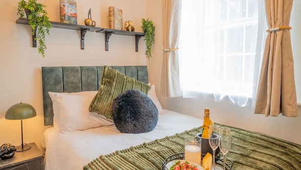 Una cama bien hecha con almohadas, una lámpara de noche, una botella de champán y un frutero en la mesita de noche.