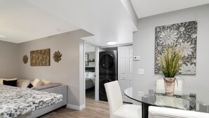 A modern bedroom with a bed, a glass dining table, and a kitchen area visible through an open door. - Lake Front - Corner Unit - 2 Bedroom 2 Bath (Brandon)