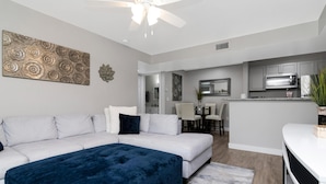 A modern living room with a white sofa, a dark blue ottoman, and a ceiling fan. - Lake Front - Corner Unit - 2 Bedroom 2 Bath (Brandon)