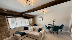 A cozy living room with a sectional sofa, a dining table with chairs, a clock on the wall, and a stone wall. - Ground floor apartment in 18th century building (Gloucestershire)