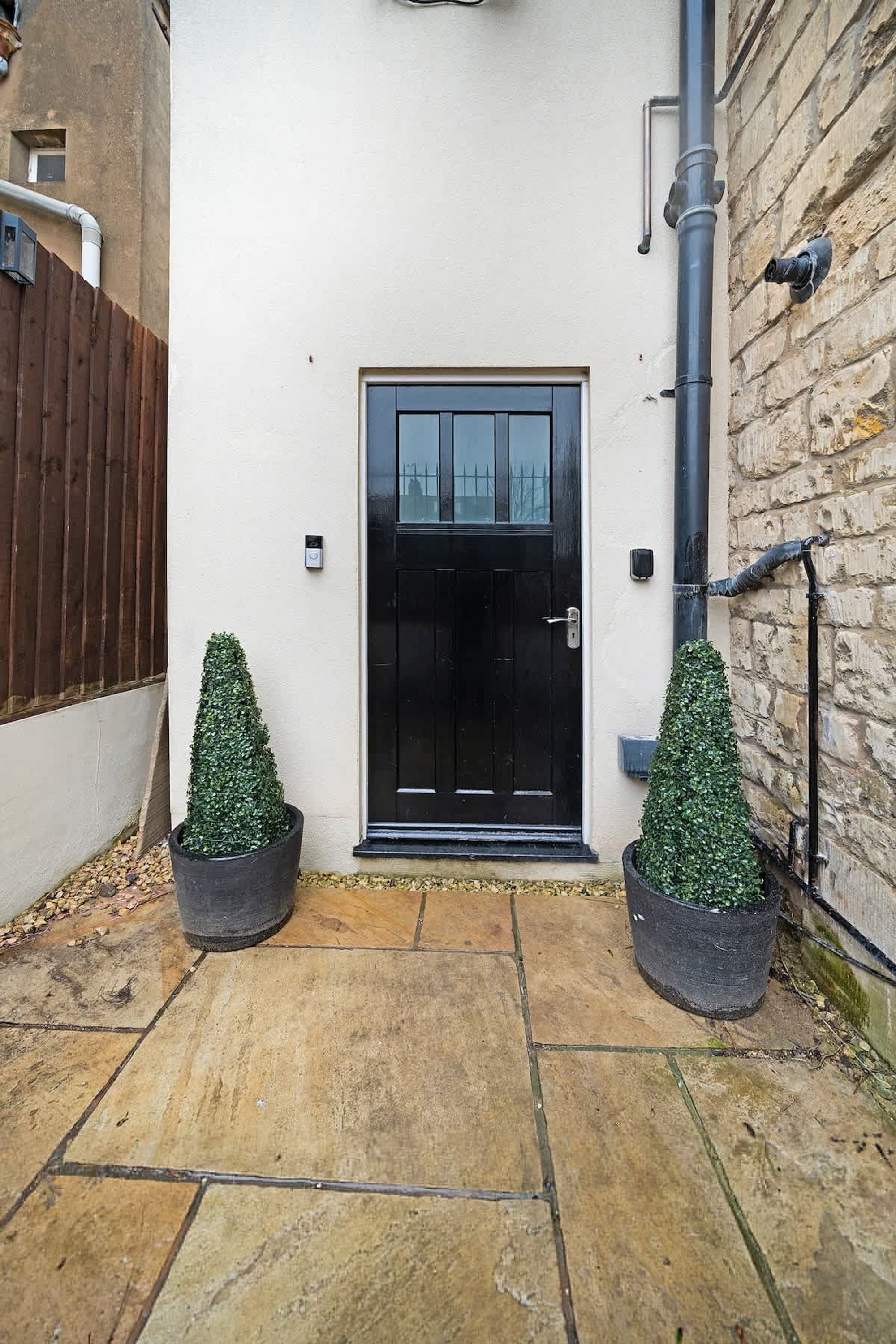 A black door with a small step leading up to it, flanked by two potted topiaries, a stone-paved entrance, and a brick wall.