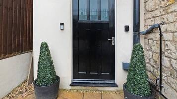 A black door with a small step leading up to it, flanked by two potted topiaries, a stone-paved entrance, and a brick wall.