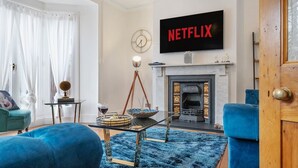 A living room with a fireplace, a television displaying 'NETFLIX,' a blue sofa, and a coffee table. - 4BD 3Bath 2Car Drive Garden Games room Nr Hospital (Gloucestershire)