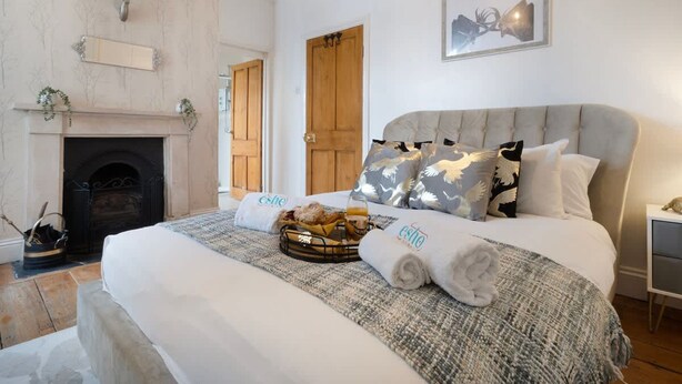 A bedroom with a fireplace, a bed with a grey headboard, and a basket of bread. - 4BD 3Bath 2Car Drive Garden Games room Nr Hospital (Gloucestershire)