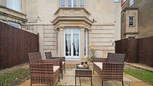 A patio with wicker furniture, a table with a wine bottle, and a building with a stone facade. - 4BD 3Bath 2Car Drive Garden Games room Nr Hospital (Gloucestershire)