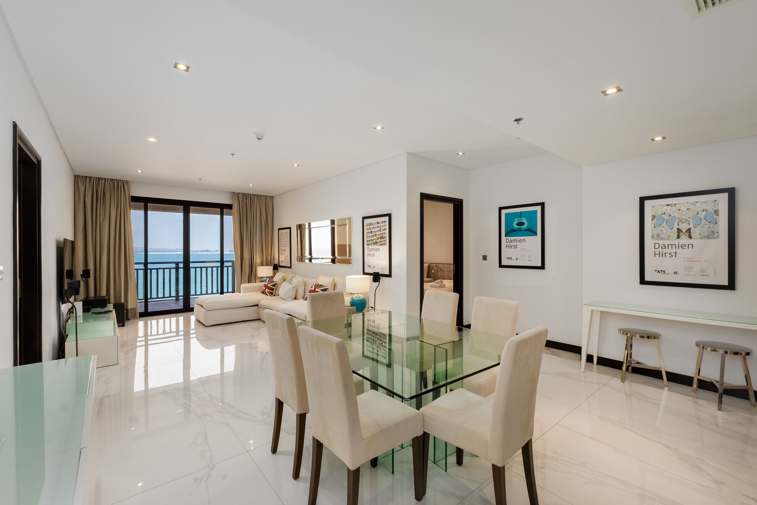 Uma sala de estar moderna com uma mesa de jantar de vidro, sofás brancos e vista para o mar.