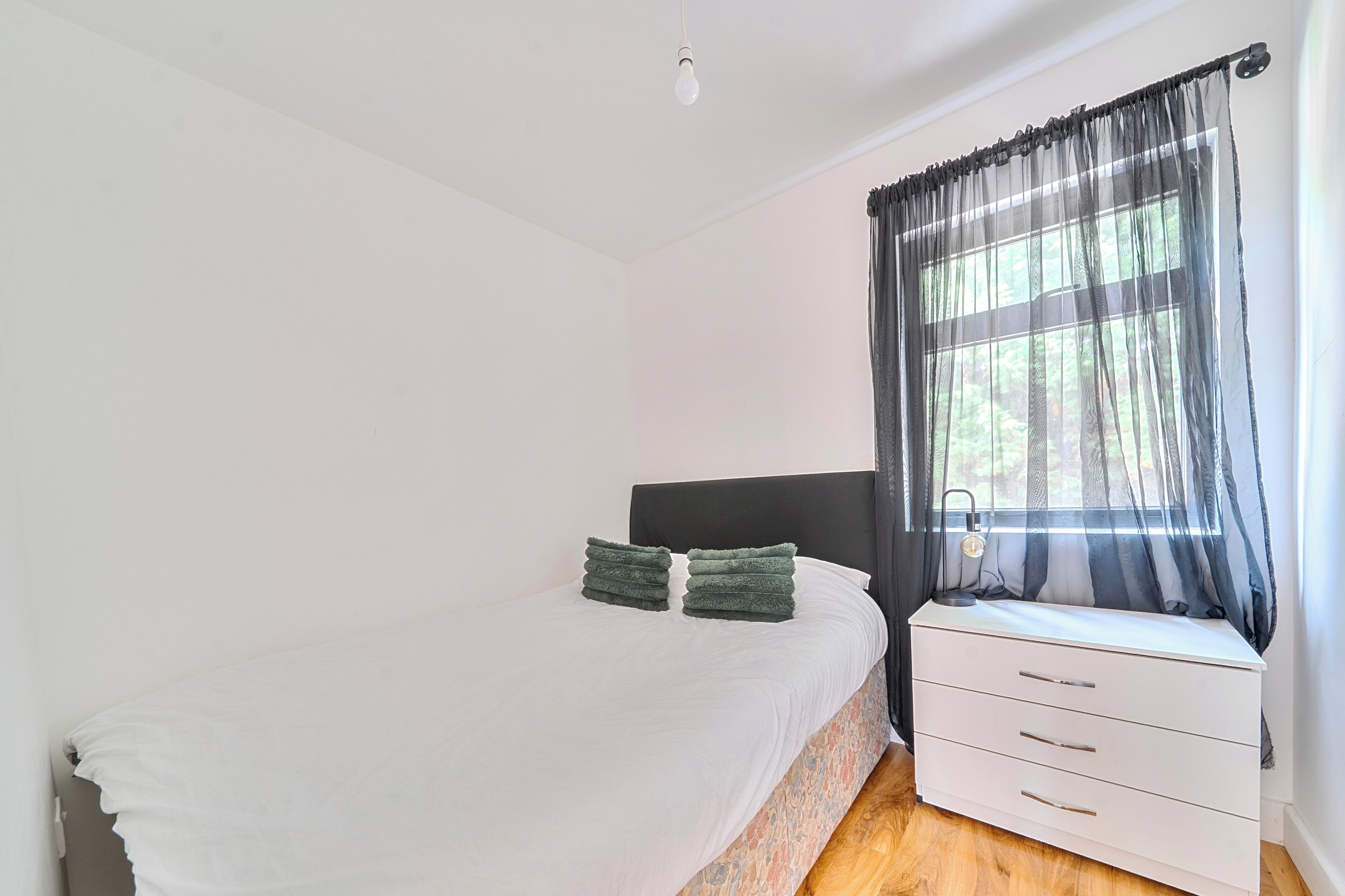 A bedroom with a bed, a bedside table, a lamp, and a window with sheer curtains.