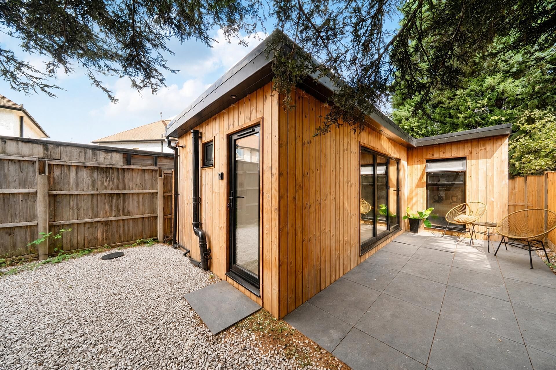 A wooden structure with a sliding door, a patio with a table and chairs, and a gravel area.