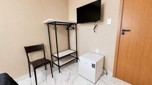 Una habitación pequeña con una silla, una estantería, un televisor montado en la pared y un electrodoméstico blanco.