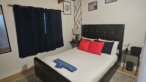 Een slaapkamer met een bed, donkere gordijnen, een hoofdbord, een nachtkastje, een lamp en een muur met ingelijste foto's.