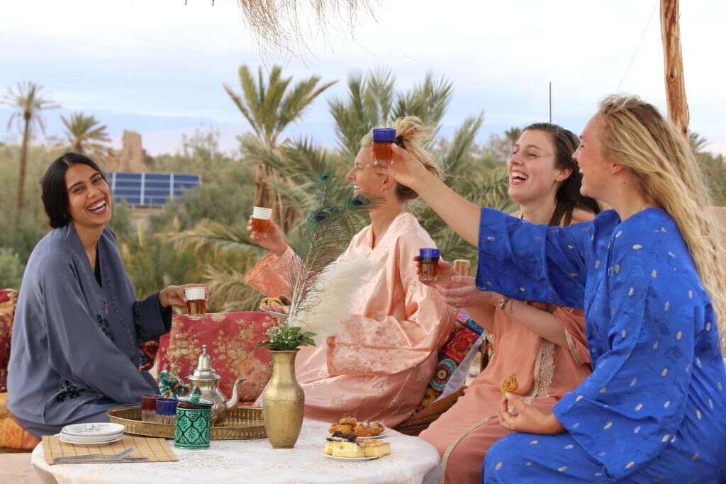 Een groep mensen zit buiten aan een tafel met een traditionele theeset, omringd door palmbomen en een heldere lucht.