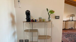 Un rincón con una estantería metálica de tres niveles, una cafetera, un florero con planta y un escritorio con cajones.