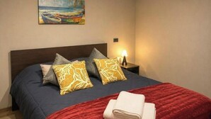 Ein ordentlich gemachtes Bett mit roter Decke und Kissen, ein Gemälde von Booten an der Wand und eine Nachttischlampe.