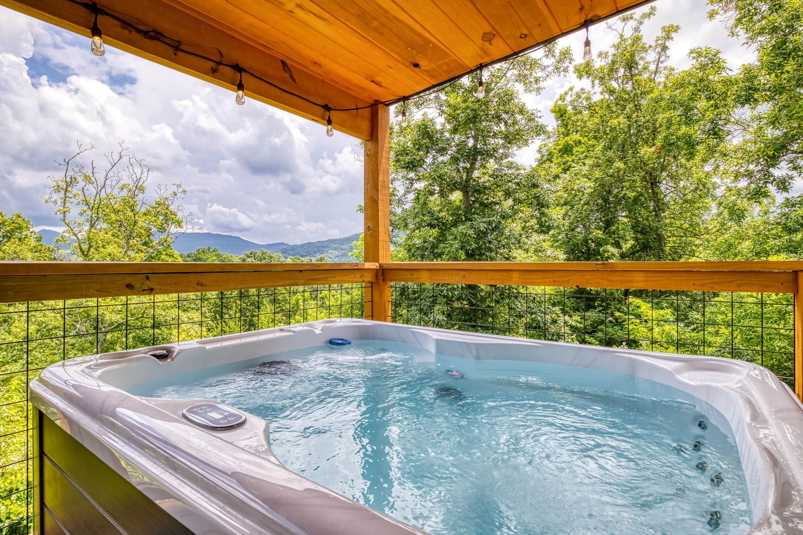 Un jacuzzi con panel de control, rodeado por una terraza de madera y barandilla metálica, con vistas a un paisaje verde exuberante con árboles y montañas a lo lejos.