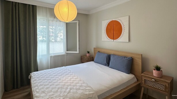 Un dormitorio con una cama, una mesita de noche, una lámpara colgante, una ventana con cortinas y una obra de arte en la pared.