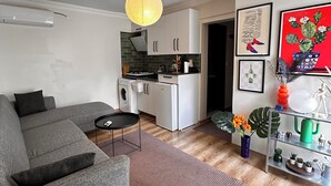Una sala moderna con un sofá gris, una mesita redonda, una zona de cocina con armarios blancos y un jarrón rojo con flores.