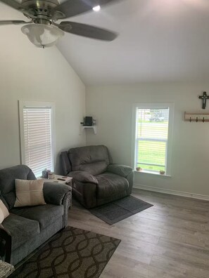 A living room with a ceiling fan, two windows with blinds, a gray sofa, and a patterned rug. - Welcome to Lilly's Pad (Aquilla)