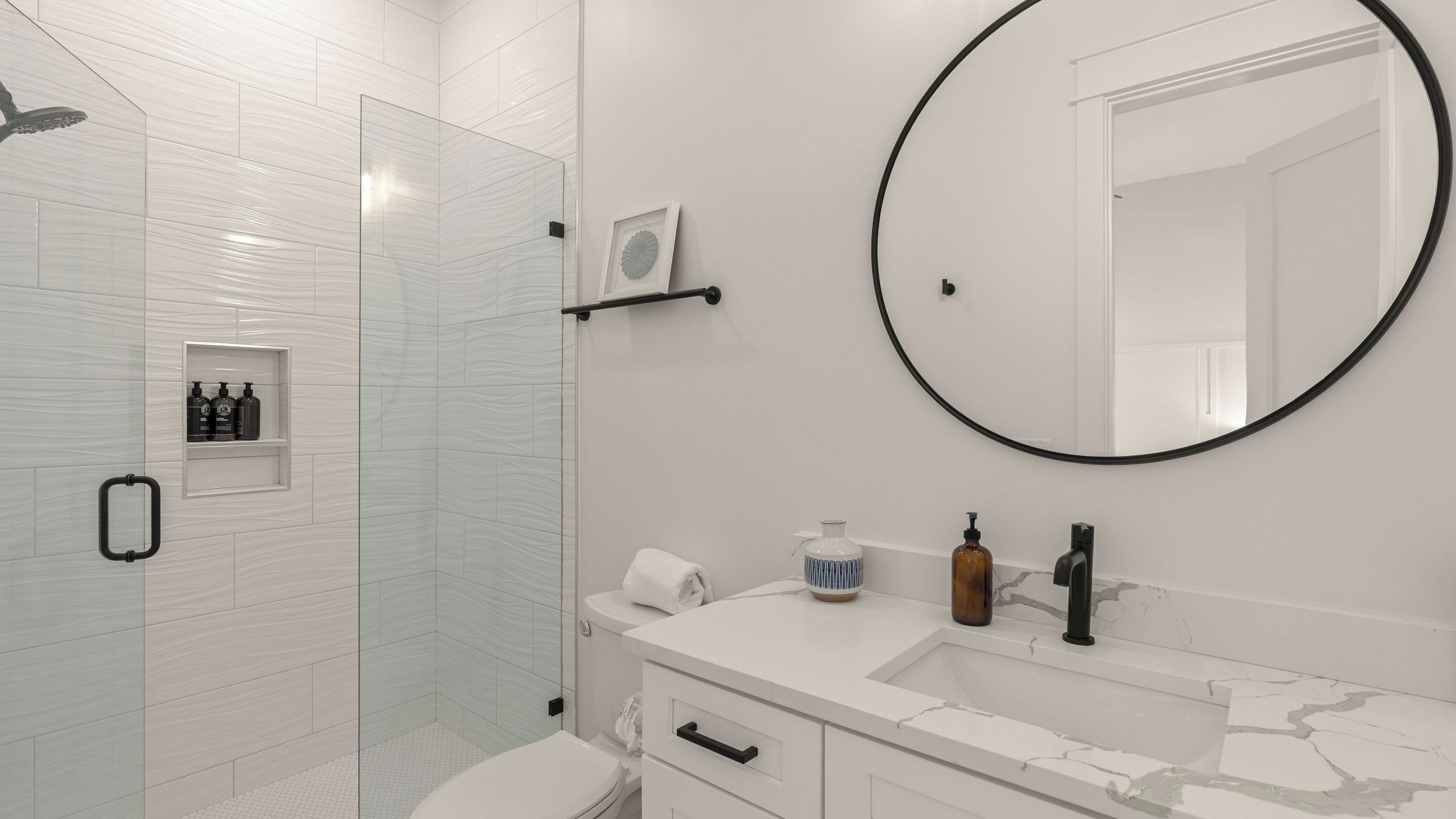 A modern bathroom with a glass shower enclosure, a round mirror, a white sink with a marble countertop, and a small shelf with toiletries.