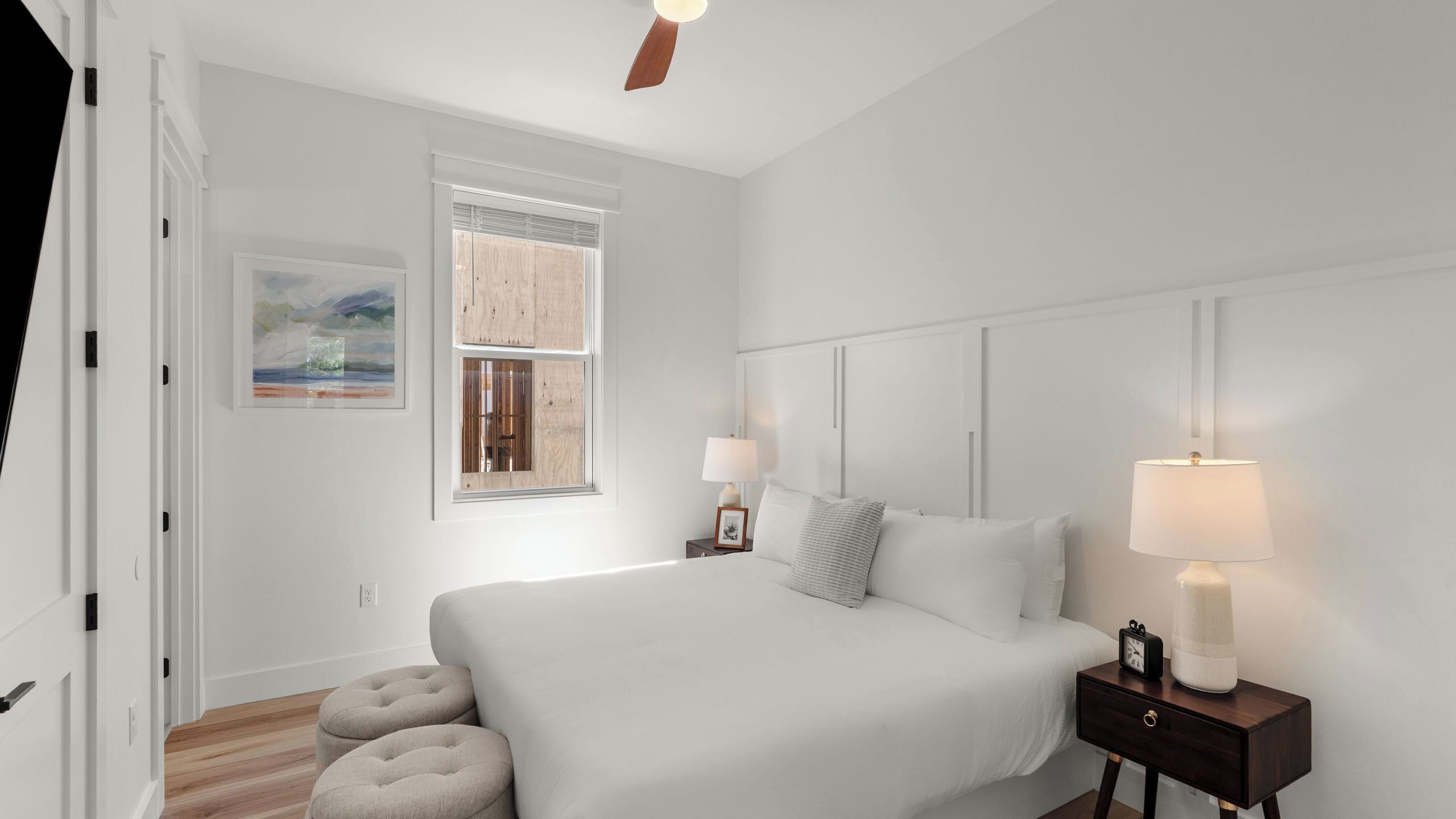 Une chambre à coucher avec un lit, des lampes de chevet, une table de nuit, une fenêtre avec des stores, un ventilateur de plafond et un tableau encadré au mur.