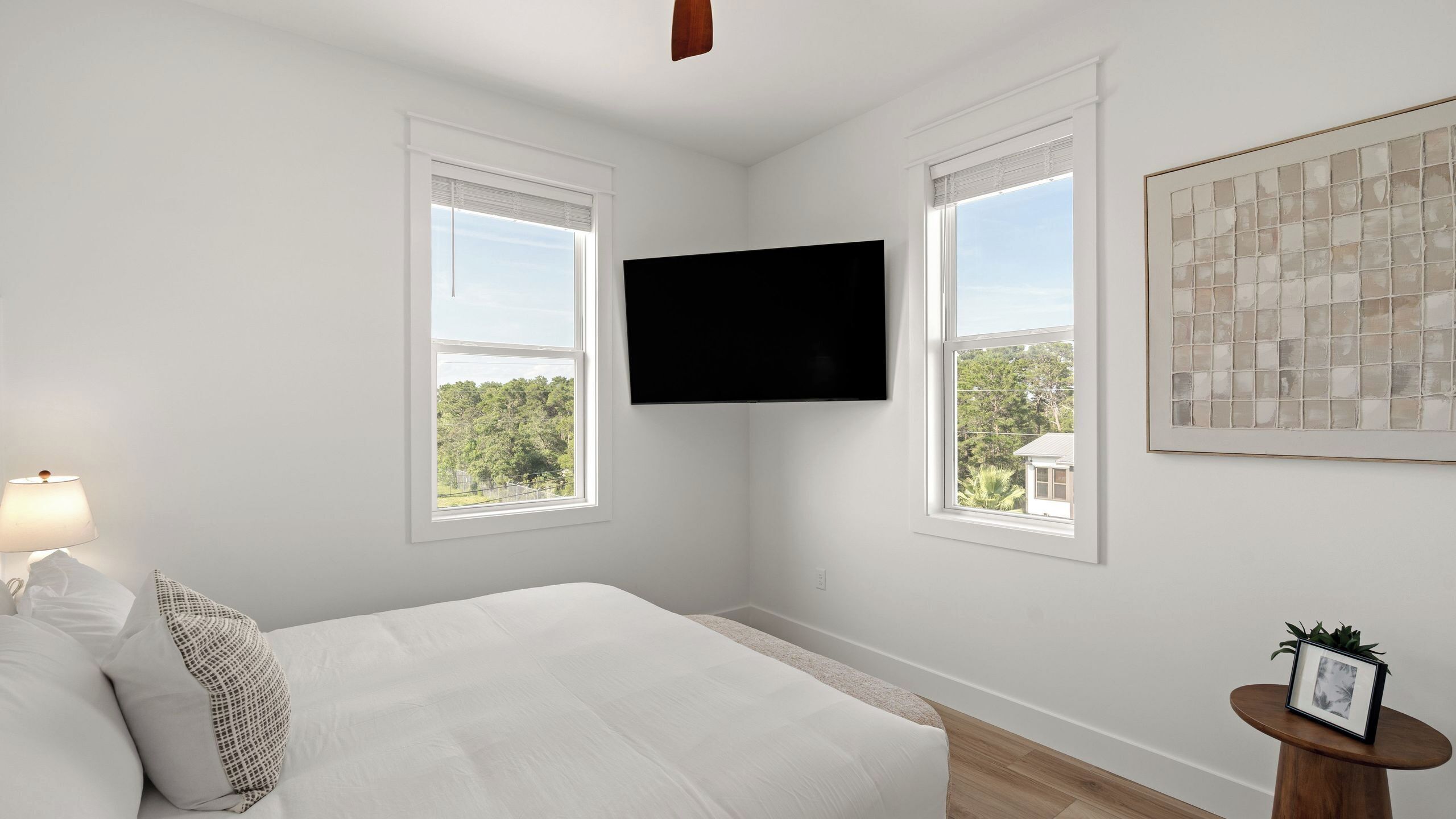 Une chambre à coucher avec un lit, deux fenêtres, un téléviseur fixé au mur et un tableau encadré accroché au mur.