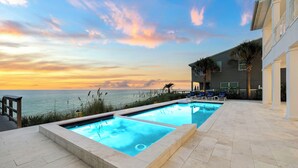 Une piscine avec vue sur l’océan au coucher du soleil, une terrasse en bois et un bâtiment doté d’un balcon.