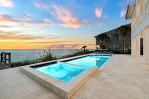 Ein Swimmingpool mit Blick auf den Ozean bei Sonnenuntergang, eine Holzterrasse und ein Gebäude mit einem Balkon.