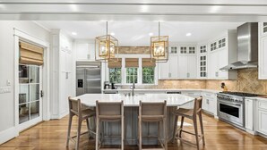 Uma cozinha moderna com armários brancos, uma ilha central com banquetas, uma coifa de aço inoxidável e luminárias pendentes.
