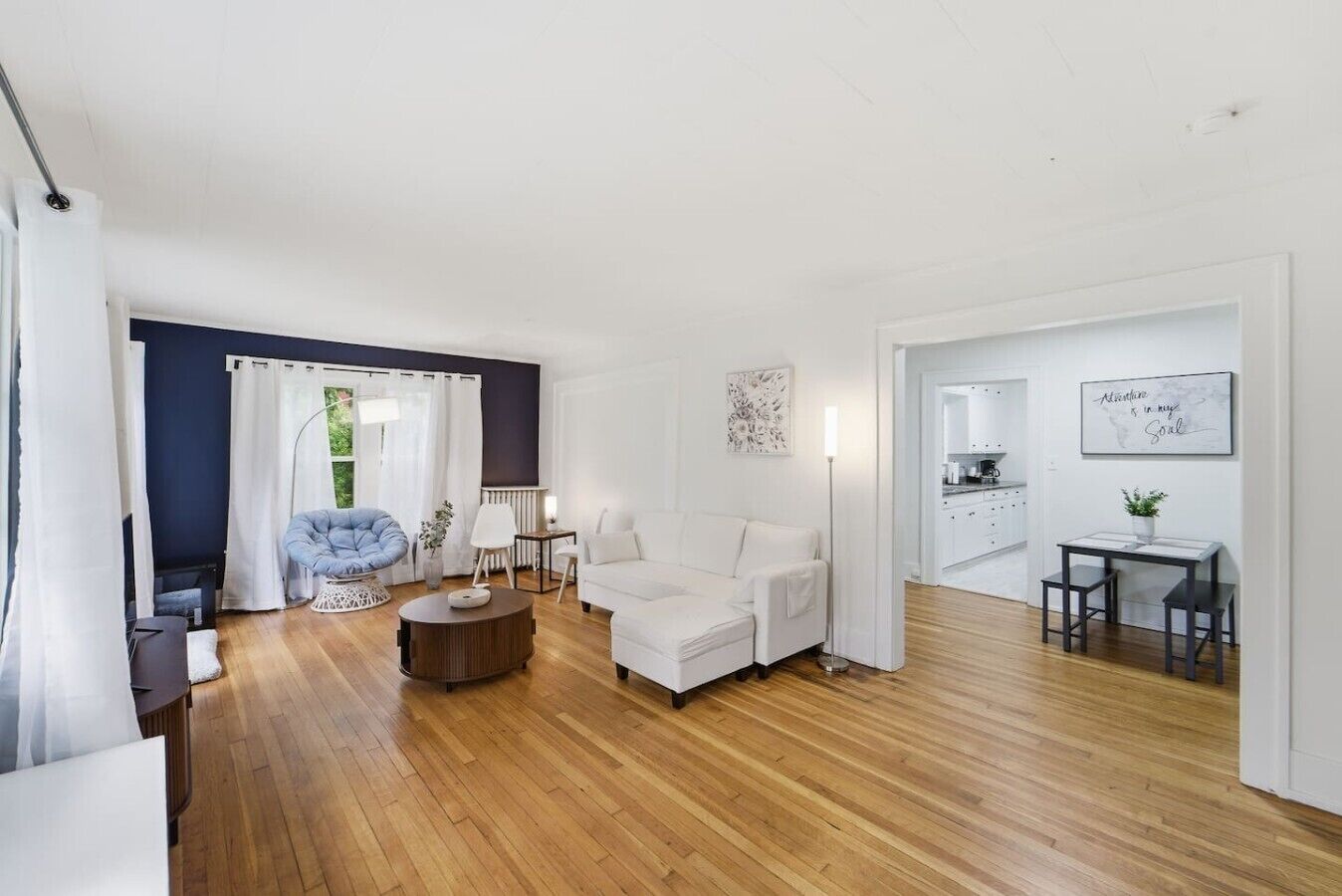 Un salon moderne avec un canapé, un fauteuil et une table basse, un coin repas avec une table et des chaises, et une cuisine visible à travers une porte ouverte.