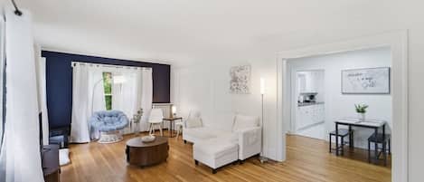 Un salon moderne avec un canapé, un fauteuil et une table basse, un coin repas avec une table et des chaises, et une cuisine visible à travers une porte ouverte.