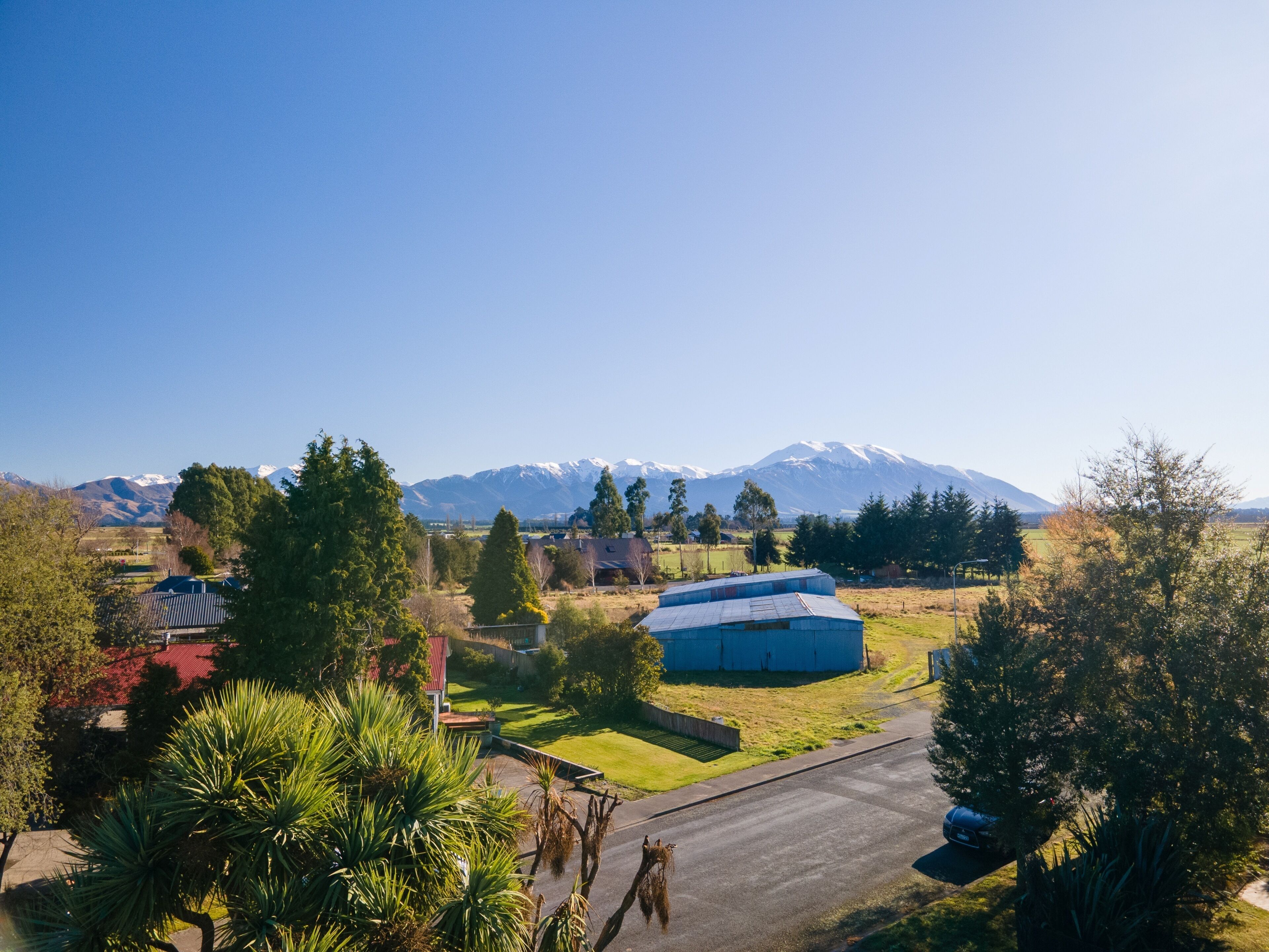 A rural landscape with a clear road, a blue building, and snow-capped mountains in the background.