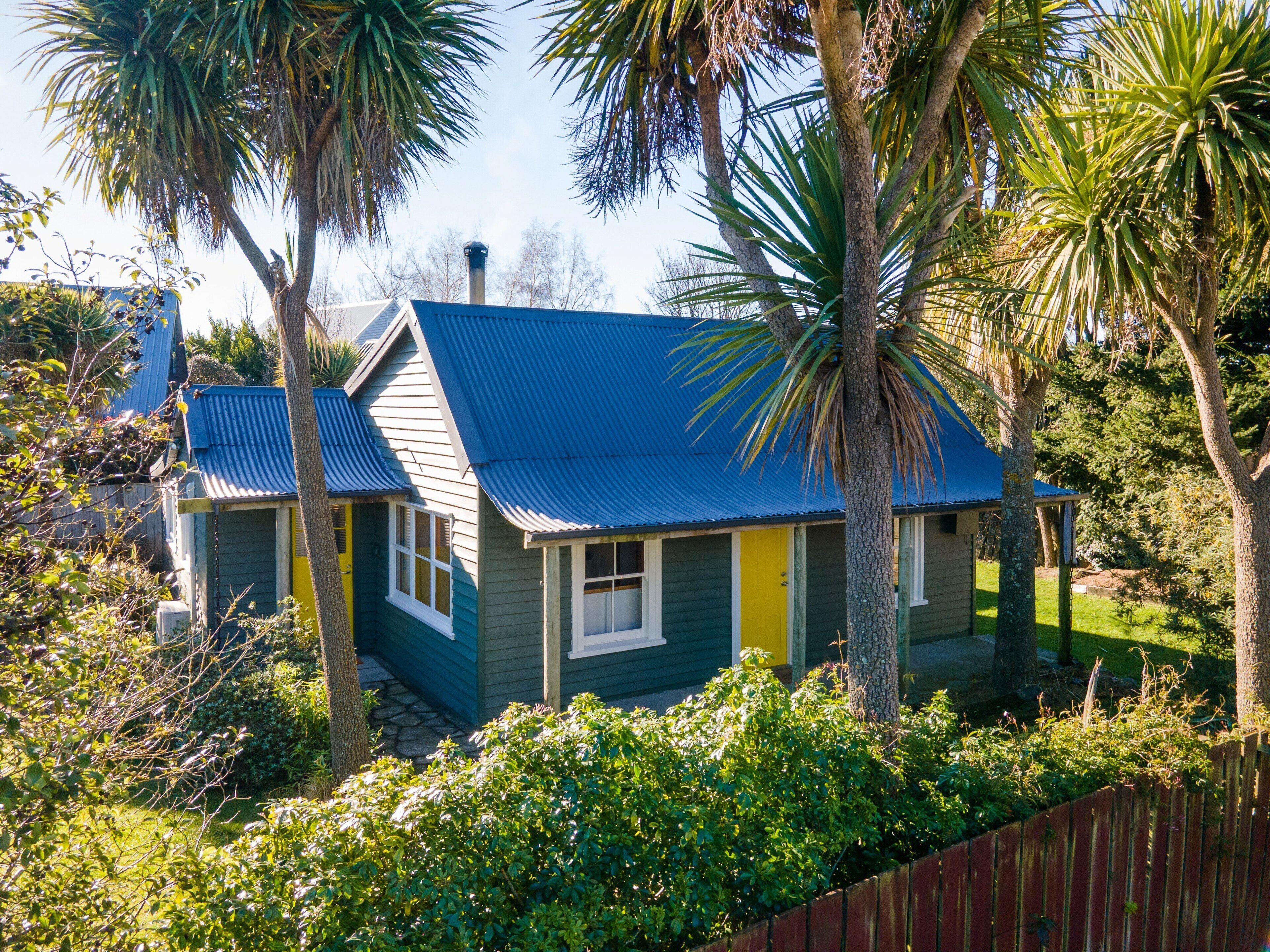 A house with a blue roof, green walls, and a yellow door, surrounded by lush greenery and palm trees.