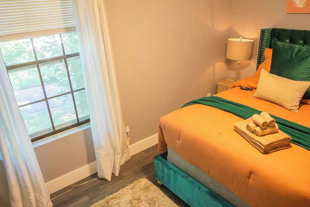Een slaapkamer met een bed, gordijnen voor het raam, een bedlampje en uitzicht op groen.